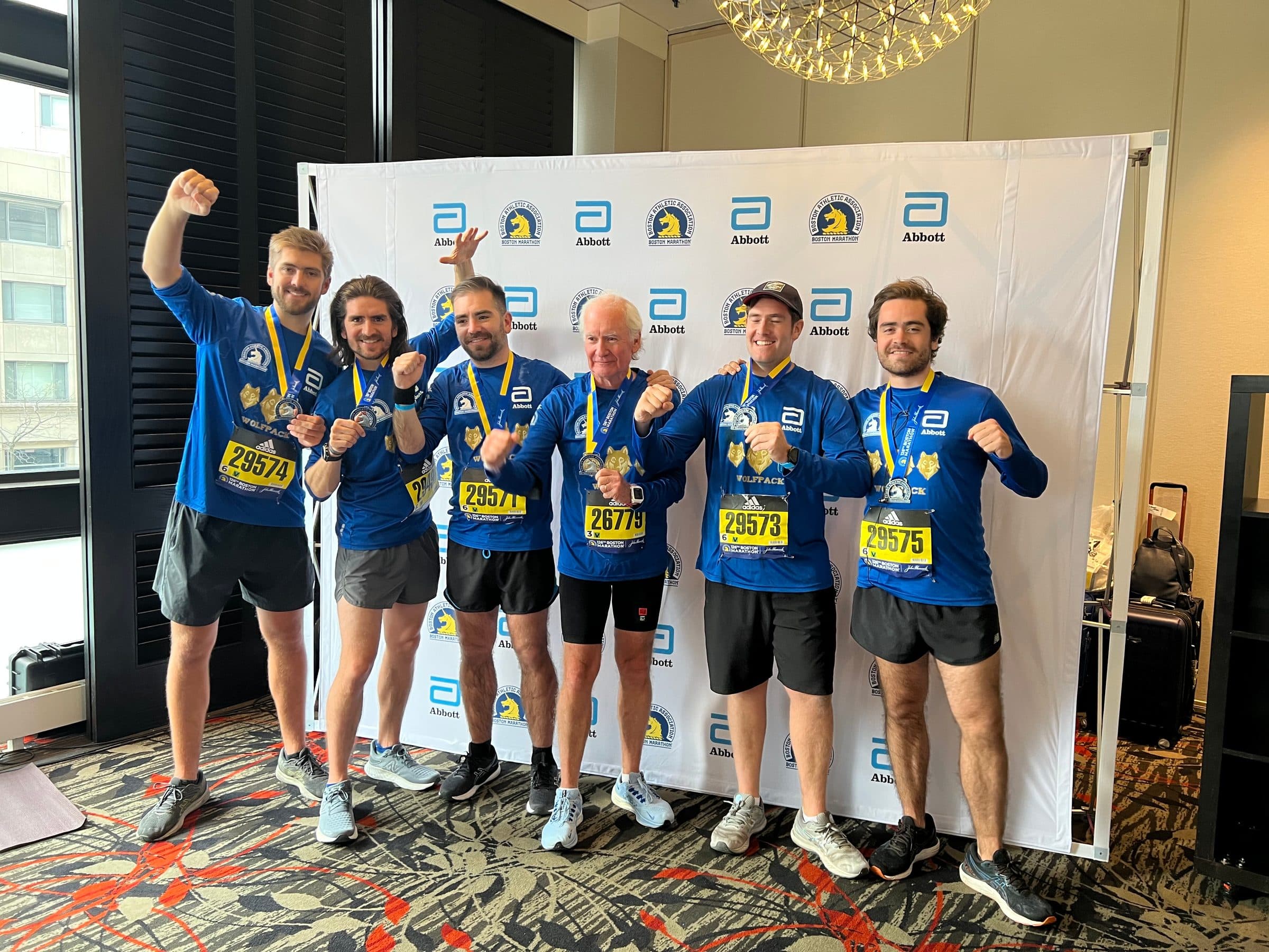 The six runners with their medals, arms raised, in front of the Abbott wall in Boston.